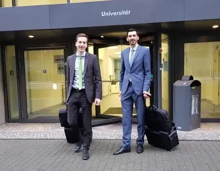 Zwei zugewandte Prüfer von BerKon stehen mit Ziehkoffern vor einer Universität.