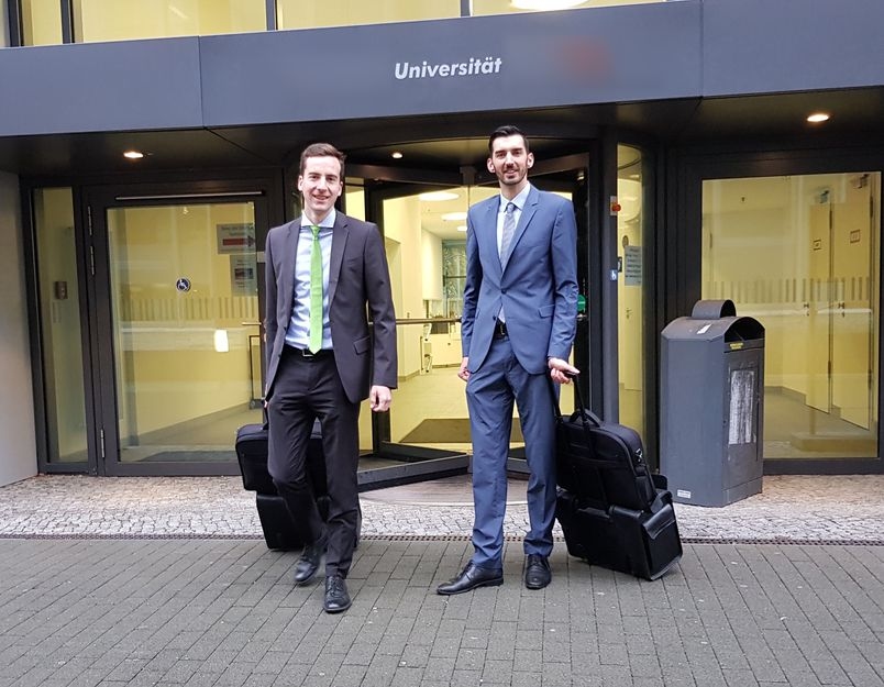Zwei zugewandte Prüfer von BerKon stehen mit Ziehkoffern vor einer Universität.